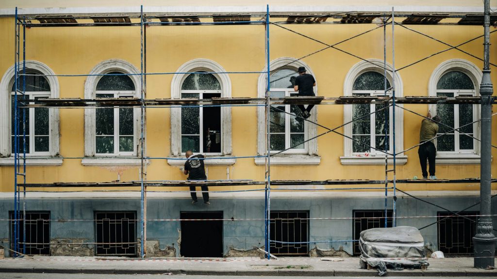 Workers on scaffolding repairing a yellow building facade.
